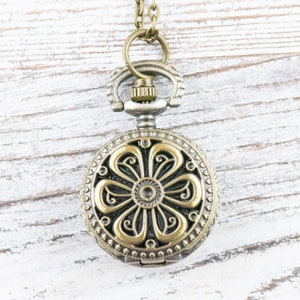 May include: A gold-toned pocket watch with a floral design on the face. The watch is on a chain and hanging from a white wooden surface.