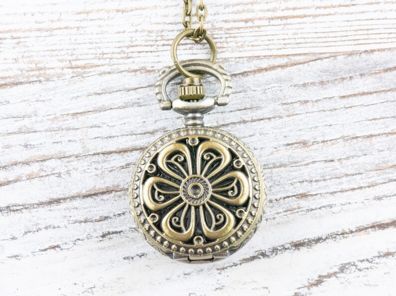 May include: A gold-toned pocket watch with a floral design on the face. The watch is on a chain and hanging from a white wooden surface.