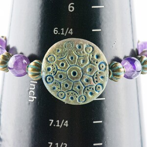 May include: Purple amethyst and gold tone bead bracelet with a round, teal and gold patterned charm. The bracelet measures 7 1/4 inches in circumference.