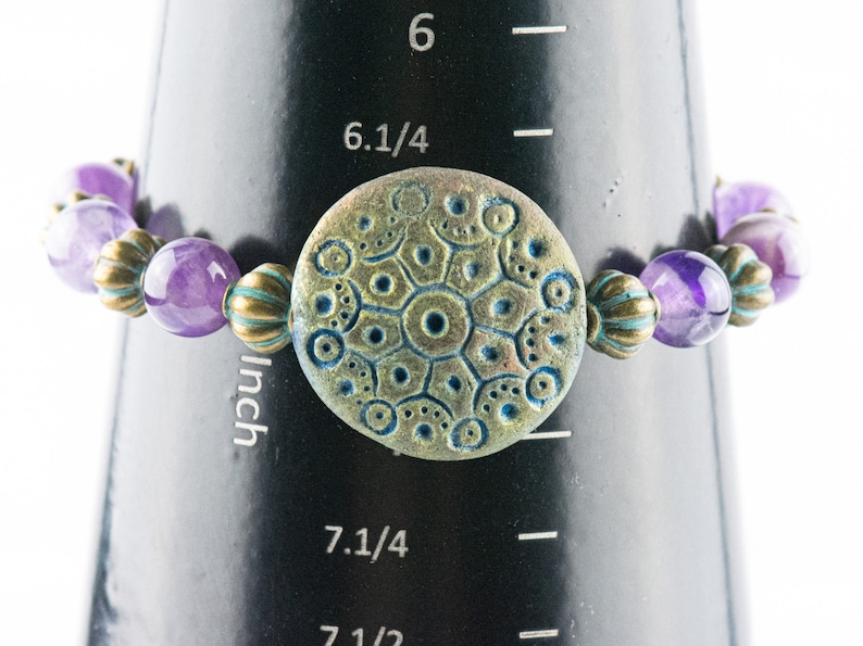 May include: Purple amethyst and gold tone bead bracelet with a round, teal and gold patterned charm. The bracelet measures 7 1/4 inches in circumference.