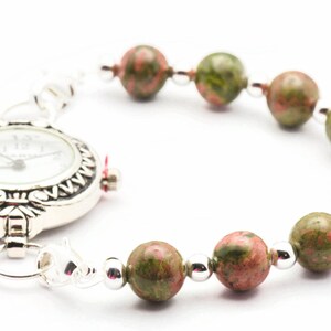 May include: A silver pocket watch with a white face and black markings is attached to a bracelet made of green and pink speckled beads.