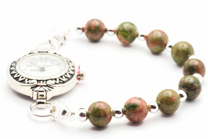 May include: A silver pocket watch with a white face and black markings is attached to a bracelet made of green and pink speckled beads.
