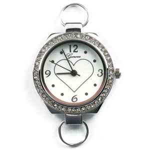 May include: A silver watch with a white face and a heart-shaped cutout. The watch is surrounded by a row of clear rhinestones. The watch has a Geneva logo on the face.