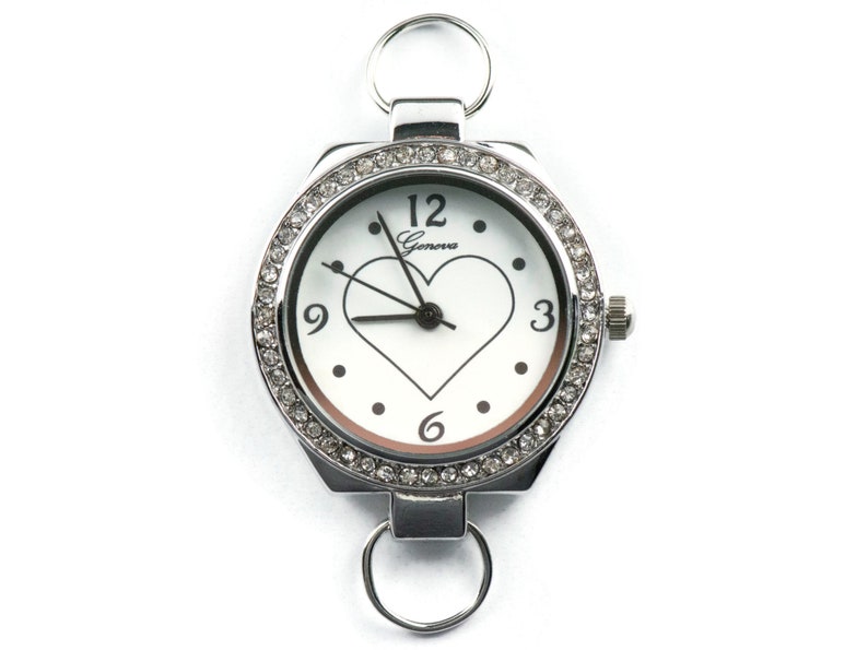 May include: A silver watch with a white face and a heart-shaped cutout. The watch is surrounded by a row of clear rhinestones. The watch has a Geneva logo on the face.