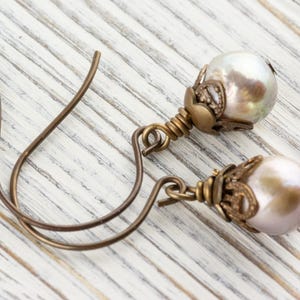 May include: A pair of dangle earrings featuring iridescent pearl-like beads. The beads are accented with ornate, antique-style metalwork and suspended from curved ear wires. The earrings rest on a white, weathered wooden surface.
