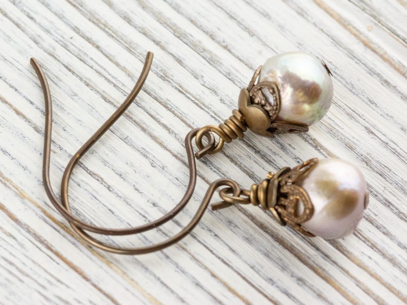 May include: A pair of dangle earrings featuring iridescent pearl-like beads. The beads are accented with ornate, antique-style metalwork and suspended from curved ear wires. The earrings rest on a white, weathered wooden surface.
