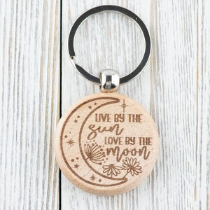 May include: Wooden keychain with a crescent moon design and the text "Live by the sun love by the moon".