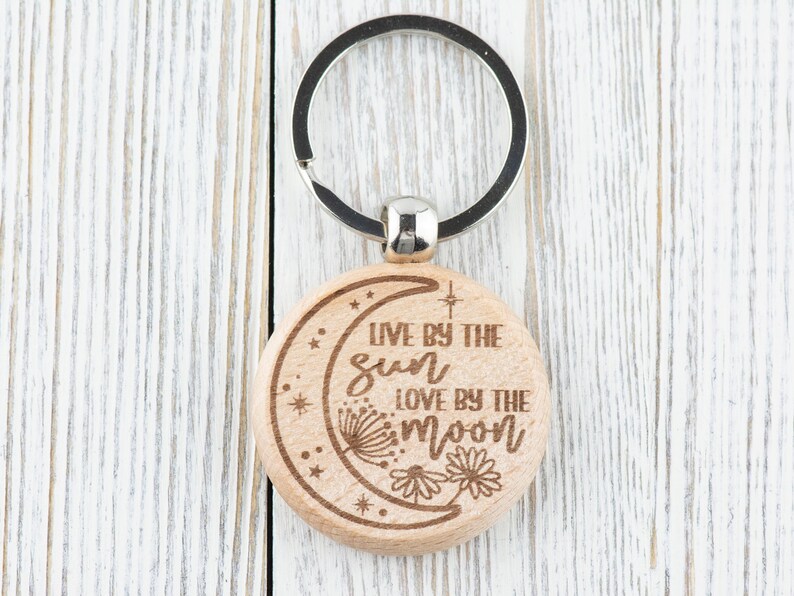 May include: Wooden keychain with a crescent moon design and the text "Live by the sun love by the moon".