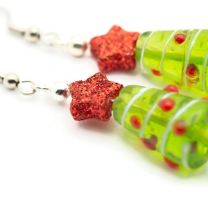 Op de afbeelding: Een paar groene glazen kerstboom oorbellen met rode glittersterren. De oorbellen zijn op een witte achtergrond.