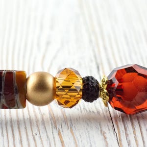Puede incluir: Primer plano de un collar de cuentas con varias formas y colores. Las cuentas incluyen una mezcla de elementos &aacute;mbar, dorados y marr&oacute;n oscuro. El collar tiene una cuenta de cristal facetada de color rojo anaranjado.