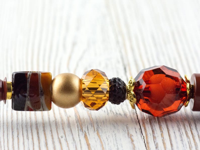 Puede incluir: Primer plano de un collar de cuentas con varias formas y colores. Las cuentas incluyen una mezcla de elementos &aacute;mbar, dorados y marr&oacute;n oscuro. El collar tiene una cuenta de cristal facetada de color rojo anaranjado.
