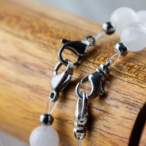 May include: Two silver clasps with clear elastic cord and white opaque beads. The clasps are on a wooden surface.
