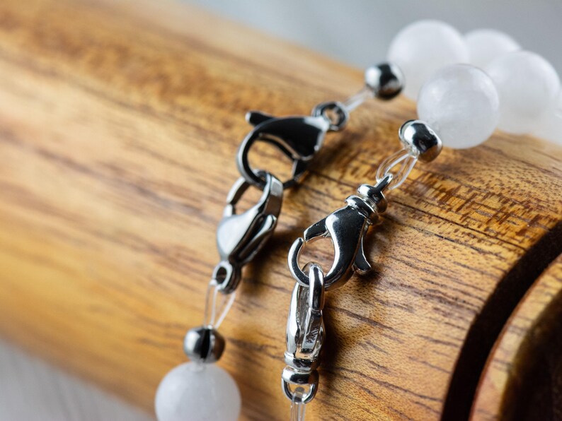 May include: Two silver clasps with clear elastic cord and white opaque beads. The clasps are on a wooden surface.