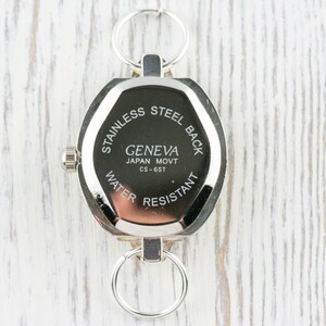Op de afbeelding: De achterkant van een zilveren horloge met een ronde wijzerplaat en een roestvrijstalen achterkant. De wijzerplaat van het horloge heeft de tekst "Geneva Japan Movt CS-6ST" en "Stainless Steel Back Water Resistant".