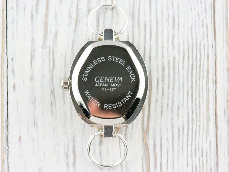 Op de afbeelding: De achterkant van een zilveren horloge met een ronde wijzerplaat en een roestvrijstalen achterkant. De wijzerplaat van het horloge heeft de tekst "Geneva Japan Movt CS-6ST" en "Stainless Steel Back Water Resistant".
