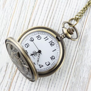 May include: A gold-toned pocket watch with a white face and black hands. The watch is open and the chain is visible. The watch is on a white wooden surface.