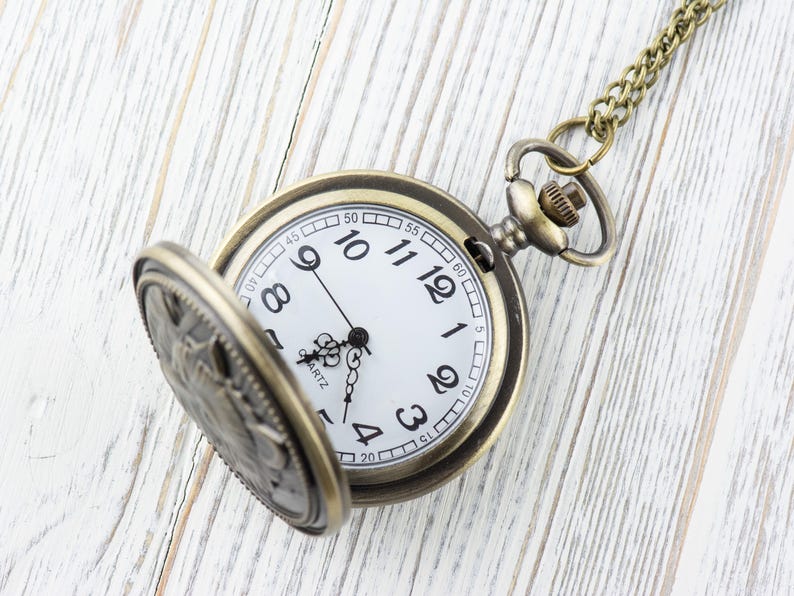 May include: A gold-toned pocket watch with a white face and black hands. The watch is open and the chain is visible. The watch is on a white wooden surface.