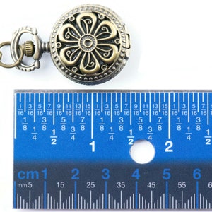 May include: A gold-toned pocket watch with a floral design on the face. The watch is attached to a chain and is being measured with a ruler. The ruler shows that the watch is approximately 1.5 inches in diameter.