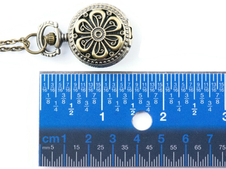 May include: A gold-toned pocket watch with a floral design on the face. The watch is attached to a chain and is being measured with a ruler. The ruler shows that the watch is approximately 1.5 inches in diameter.