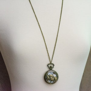 Puede incluir: Collar con colgante de reloj de bolsillo antiguo en tono bronce. La esfera redonda del reloj presenta una ilustraci&oacute;n detallada de un conejo y un reloj. El reloj cuelga de una larga cadena.