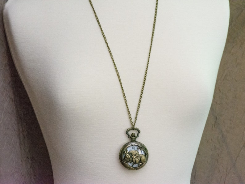 Puede incluir: Collar con colgante de reloj de bolsillo antiguo en tono bronce. La esfera redonda del reloj presenta una ilustraci&oacute;n detallada de un conejo y un reloj. El reloj cuelga de una larga cadena.