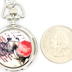 Puede incluir: Reloj de bolsillo plateado con una esfera blanca que presenta un dise&ntilde;o de postal vintage con una bicicleta, una rosa y texto en franc&eacute;s. El reloj es aproximadamente del tama&ntilde;o de una moneda de 25 c&eacute;ntimos de euro.