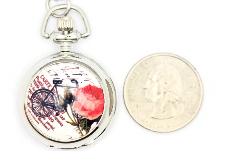 Puede incluir: Reloj de bolsillo plateado con una esfera blanca que presenta un dise&ntilde;o de postal vintage con una bicicleta, una rosa y texto en franc&eacute;s. El reloj es aproximadamente del tama&ntilde;o de una moneda de 25 c&eacute;ntimos de euro.