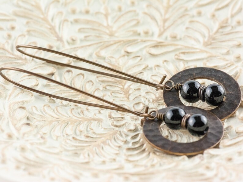Peut inclure: Une paire de boucles d'oreilles pendantes avec des pendentifs ovales en bronze martel&eacute;, chacune avec deux perles noires. Les boucles d'oreilles ont de longs et fins fils d'oreille en bronze. Elles sont pr&eacute;sent&eacute;es sur une surface blanche textur&eacute;e.