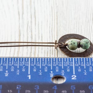 Puede incluir: Un pendiente artesanal de color bronce con un dise&ntilde;o ovalado martillado, con dos cuentas moteadas verdes y marrones. El pendiente mide aproximadamente 5 cm de largo, seg&uacute;n la regla de la imagen.