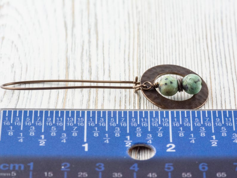 Puede incluir: Un pendiente artesanal de color bronce con un dise&ntilde;o ovalado martillado, con dos cuentas moteadas verdes y marrones. El pendiente mide aproximadamente 5 cm de largo, seg&uacute;n la regla de la imagen.