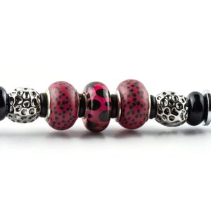 May include: A close-up of a bracelet with decorative beads. The bracelet features a mix of black, silver, and red beads. The red beads have a black polka dot pattern. The bracelet has a silver clasp on each end.