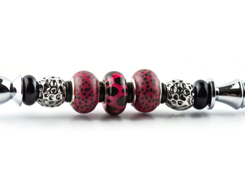 May include: A close-up of a bracelet with decorative beads. The bracelet features a mix of black, silver, and red beads. The red beads have a black polka dot pattern. The bracelet has a silver clasp on each end.