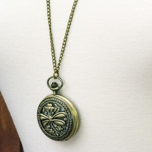 May include: A vintage-style pocket watch pendant necklace. The round, antique brass-toned watch features an ornate floral and bow design on the face. The watch hangs from a matching chain, creating a classic and elegant accessory.