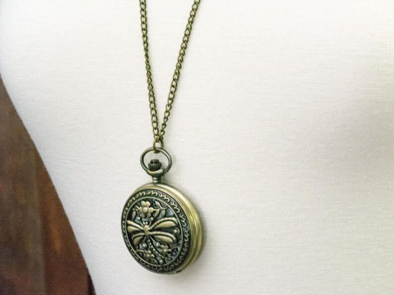 May include: A vintage-style pocket watch pendant necklace. The round, antique brass-toned watch features an ornate floral and bow design on the face. The watch hangs from a matching chain, creating a classic and elegant accessory.