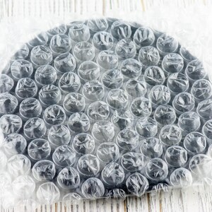 May include: A round, black object is wrapped in clear bubble wrap. The bubble wrap is partially covering the object, which is sitting on a white wooden surface. The bubbles are clear and round, and the object is a solid black color.