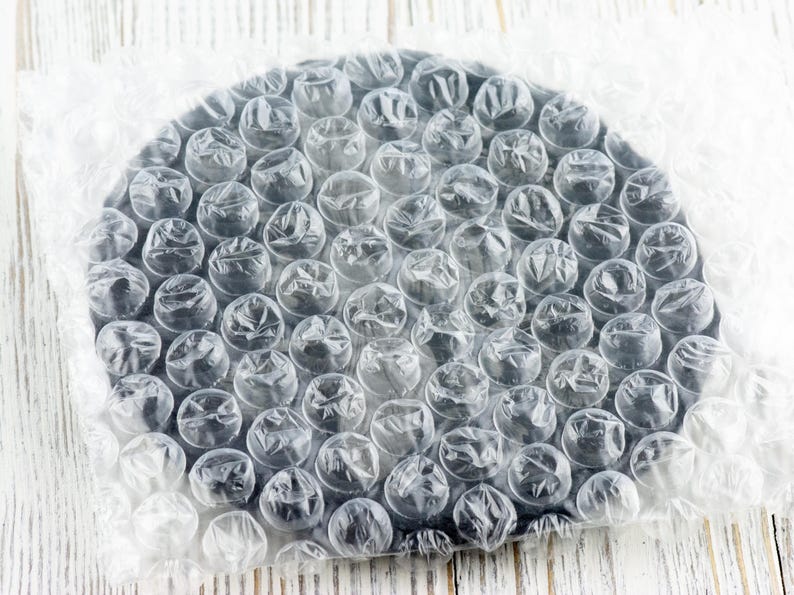 May include: A round, black object is wrapped in clear bubble wrap. The bubble wrap is partially covering the object, which is sitting on a white wooden surface. The bubbles are clear and round, and the object is a solid black color.