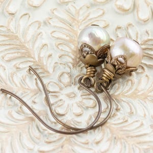 May include: A pair of pearl earrings with a vintage aesthetic. Each earring features a large, lustrous pearl, accented with ornate, antique-gold-colored metalwork. The ear wires are a simple, curved design. The earrings rest on a cream-colored, textured surface.