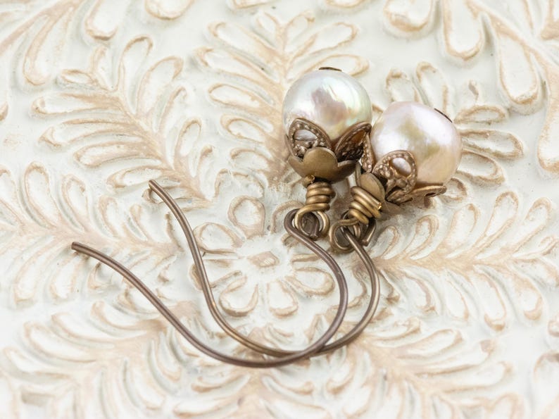 May include: A pair of pearl earrings with a vintage aesthetic. Each earring features a large, lustrous pearl, accented with ornate, antique-gold-colored metalwork. The ear wires are a simple, curved design. The earrings rest on a cream-colored, textured surface.
