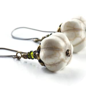 May include: A pair of white, textured, teardrop-shaped earrings with green glass beads and bronze accents. The earrings are suspended from bronze wire hooks.