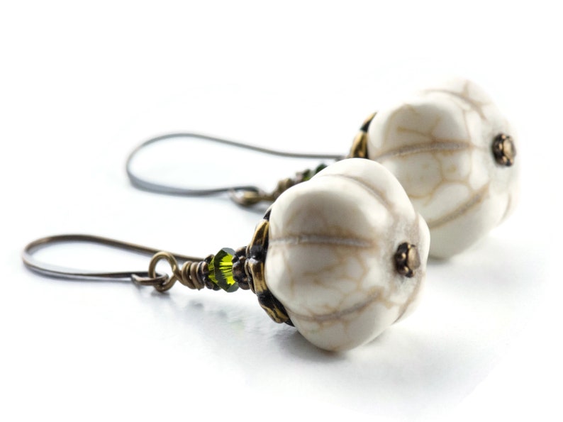 May include: A pair of white, textured, teardrop-shaped earrings with green glass beads and bronze accents. The earrings are suspended from bronze wire hooks.
