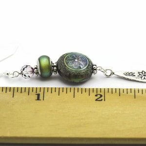 May include: A pair of dangle earrings with a silver hook, a purple crystal bead, a green iridescent bead, a green and purple patterned bead, and a silver charm with a flower design. The earrings are measured against a ruler, showing they are approximately 5 cm long.