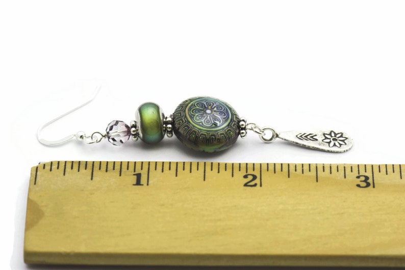 May include: A pair of dangle earrings with a silver hook, a purple crystal bead, a green iridescent bead, a green and purple patterned bead, and a silver charm with a flower design. The earrings are measured against a ruler, showing they are approximately 5 cm long.