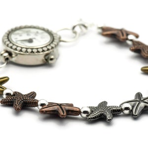 May include: A silver watch with a white face and black hands is attached to a bracelet with silver and bronze starfish charms.
