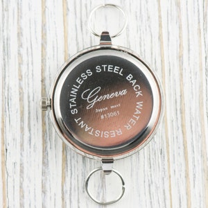Puede incluir: Un reloj de bolsillo redondo de tono plateado con una esfera central de color marr&oacute;n oscuro. La carcasa trasera tiene grabado el texto Geneva, Japan movt, #13061, Stainless Steel Back y Water Resistant. Incluye una corona y dos anillos plateados.
