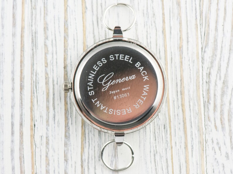 Puede incluir: Un reloj de bolsillo redondo de tono plateado con una esfera central de color marr&oacute;n oscuro. La carcasa trasera tiene grabado el texto Geneva, Japan movt, #13061, Stainless Steel Back y Water Resistant. Incluye una corona y dos anillos plateados.