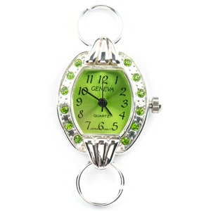 Op de afbeelding: Een zilveren horloge met een groene wijzerplaat en een zilveren band. Het horloge heeft een ronde wijzerplaat met een witte rand en groene cijfers. Het horloge is versierd met groene strasssteentjes rond de wijzerplaat. De wijzerplaat van het horloge zegt "GENEVA" en "QUARTZ".