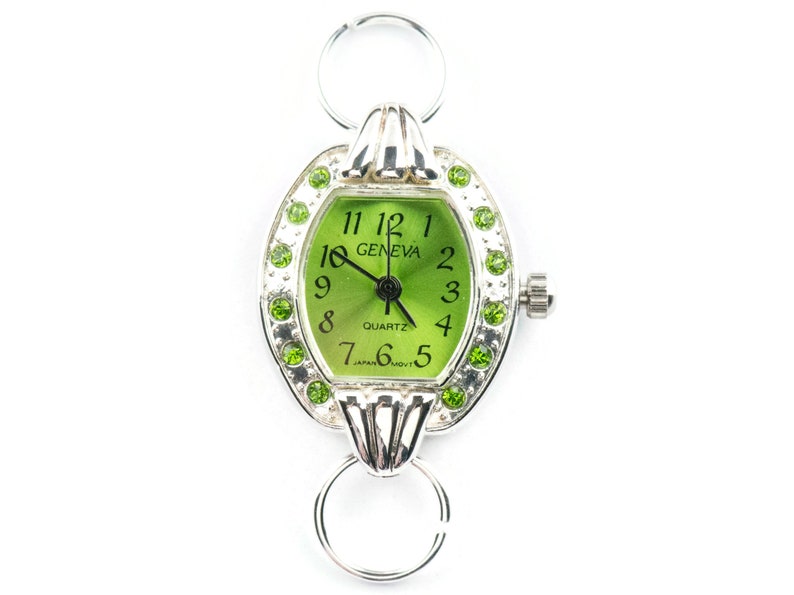Op de afbeelding: Een zilveren horloge met een groene wijzerplaat en een zilveren band. Het horloge heeft een ronde wijzerplaat met een witte rand en groene cijfers. Het horloge is versierd met groene strasssteentjes rond de wijzerplaat. De wijzerplaat van het horloge zegt "GENEVA" en "QUARTZ".