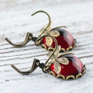 Pode incluir: Um par de brincos pendentes com caboch&otilde;es de vidro vermelho e detalhes de lib&eacute;lula dourados.