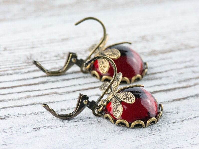 Pode incluir: Um par de brincos pendentes com caboch&otilde;es de vidro vermelho e detalhes de lib&eacute;lula dourados.