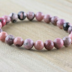 May include: A pink and gray gemstone bracelet with round beads. The bracelet is made of natural stones and is approximately 7 inches in length.